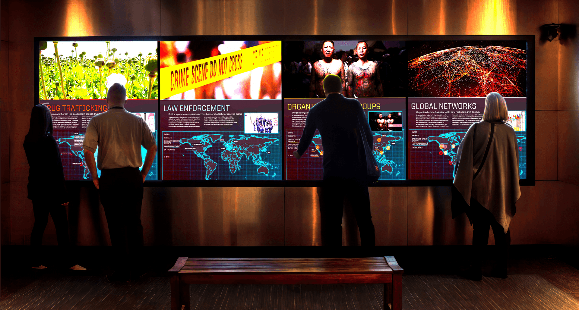 Four museum visitors stand in front of a large video screen exhibit. The screens display text and maps related to topics in global organized crime, including Drug Trafficking, Law Enforcement and Global Networks. Two of the visitors are touching the screens to make selections.
