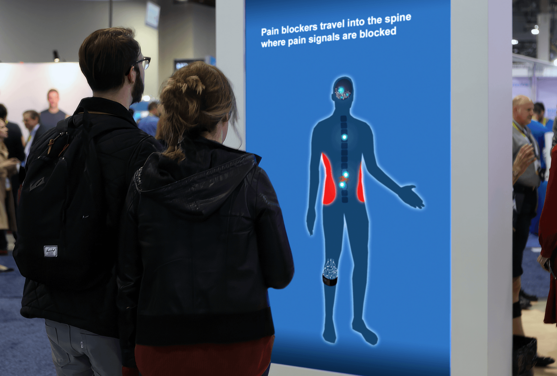 At a trade show two individuals stand in front of 90″ screen featuring the Quell Interactive. On the screen is a silhouette with highlighted areas showing pain areas and signals traveling up the spine.