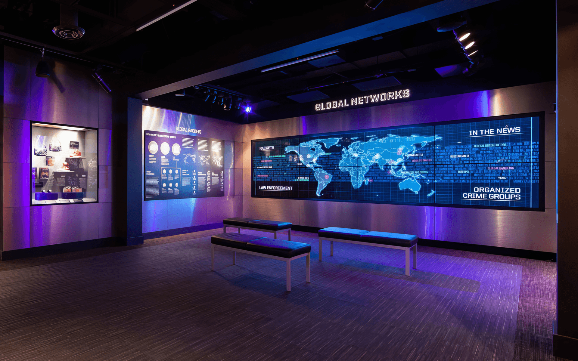 An exhibit room at The Mob Museum. Shiny metallic walls reflect dim blue and purple lighting. Three benches are arranged in front of a large video screen displaying a world map, with the title Global Networks above it on the wall. A smaller screen to the left is titled Global Rackets and displays charts and text