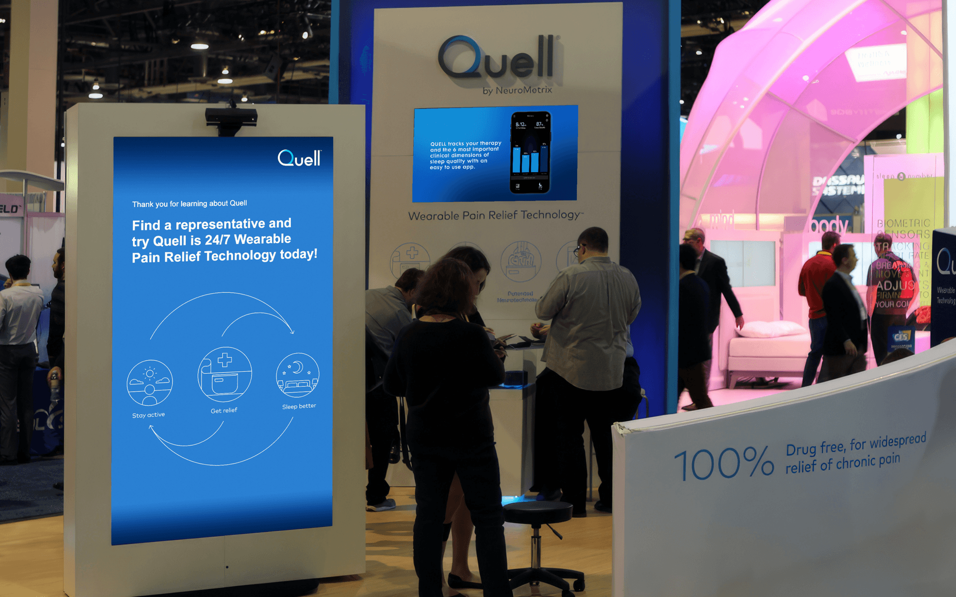 The Quell booth at CES. In the foreground a large display with a device mounted at the top thanks users for learning and directs users to speak to a representative. In the background business people talk in front of another wall mounted screen.
